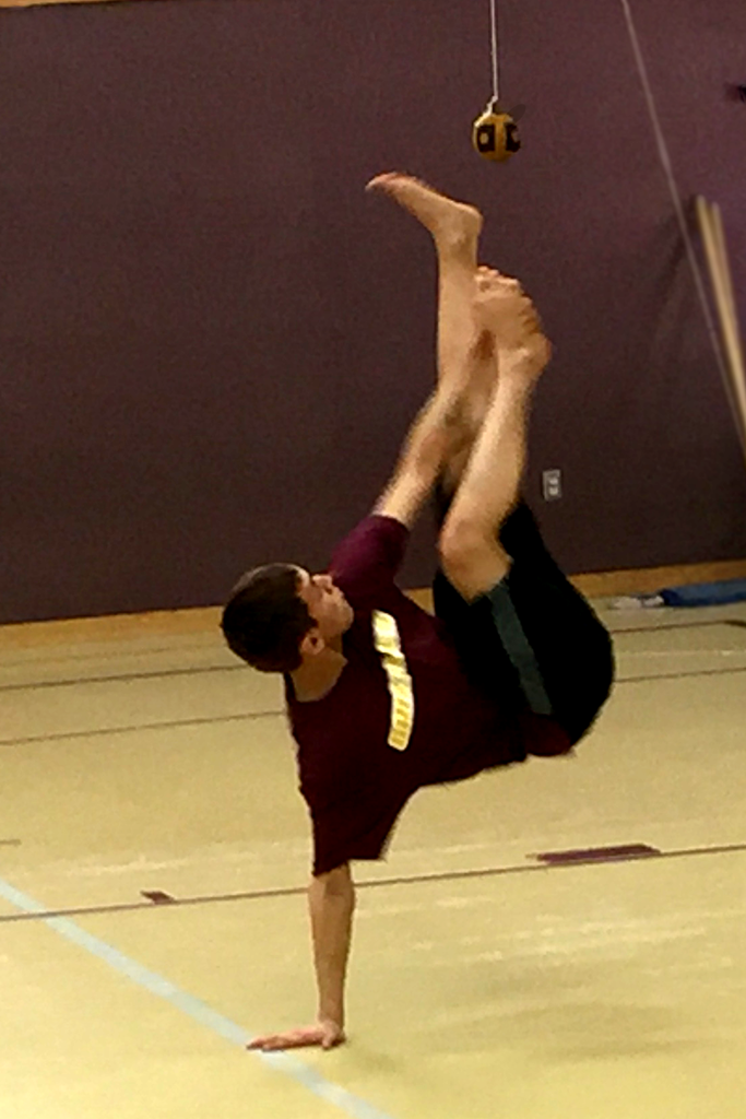 A young man balancing on his right palm while holding his right foot in his left hand and kicking a ball suspended 6 feet in the air with his left foot.